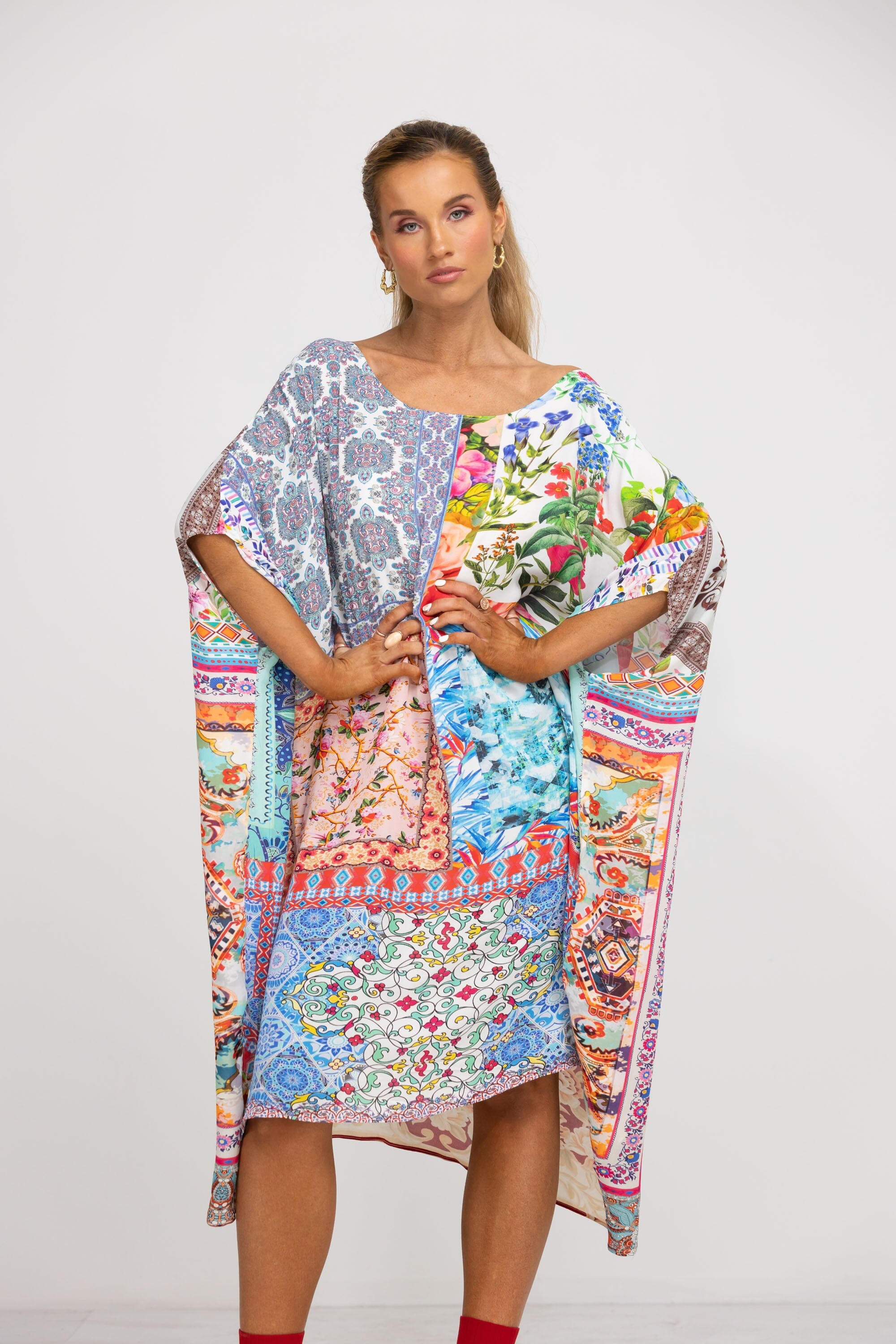 Full-front view of a model in a loose-fitting, asymmetrical kaftan dress. The garment is midi-length, with one exposed shoulder, and covered in a highly detailed, colorful, tiled patchwork pattern.
