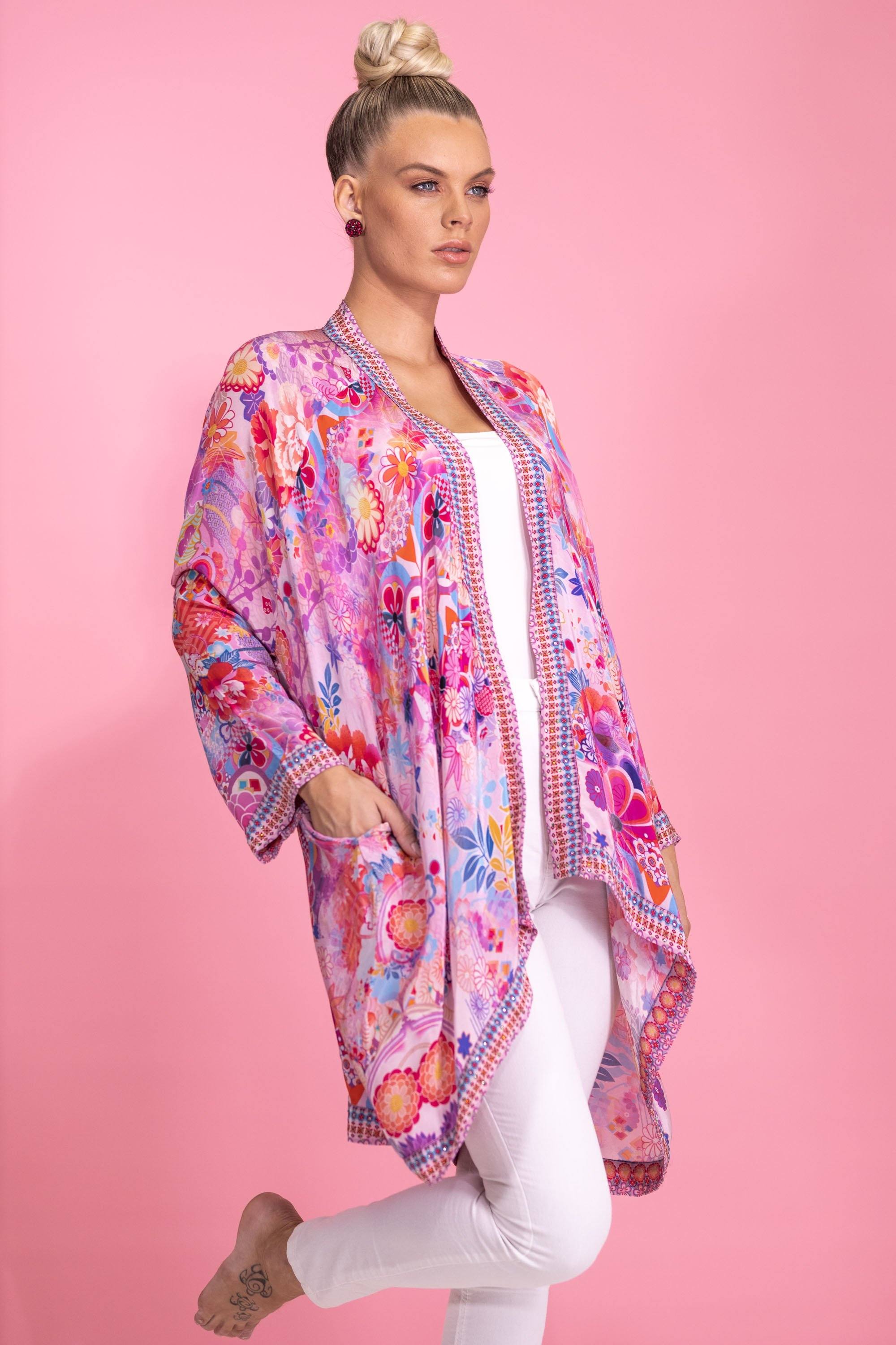 Bondi (TheSwankStore) floral kimono cardigan. A woman models an open-front, long-line kimono-style cardigan featuring an all-over vibrant pink, orange, and purple floral print with intricate border details. Worn over a white top and pants against a pink background.
