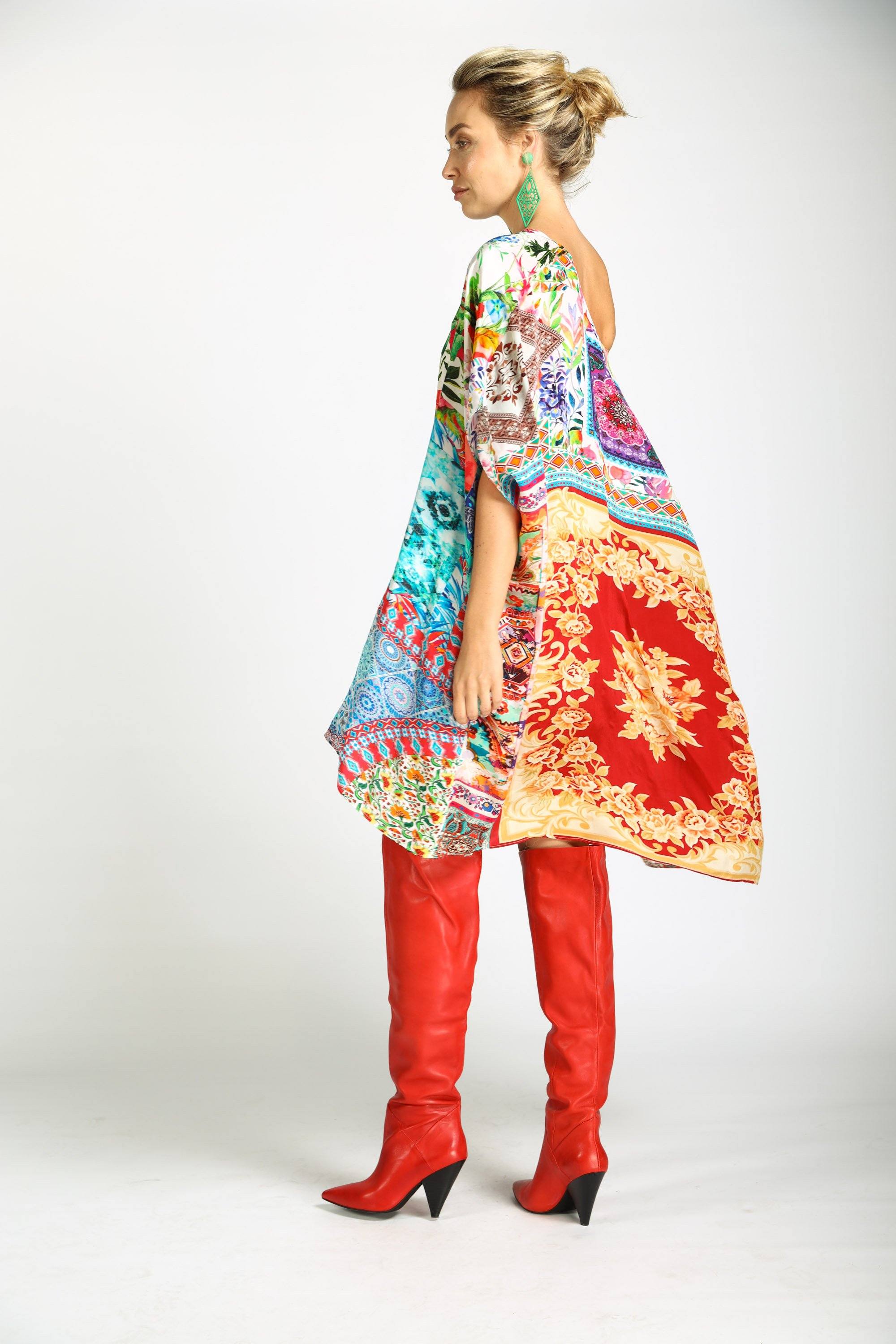 Side view with flow of the Bohemian-style Short Box Kaftan, Damascus print. The kaftan dress is worn with red knee-high boots, highlighting the asymmetrical, flowing hemline and the vibrant mix of blue, red, pink, and white prints.